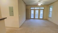 This single-floor space offers a welcoming and functional layout designed for comfortable everyday living. The main living area is a standout feature, characterized by plush wall-to-wall carpeting that adds warmth underfoot and a sense of coziness throughout. A ceiling fan with a light fixture is centrally mounted, providing both air circulation and ambient lighting to keep the space comfortable year-round. Natural light is a defining element of this room, streaming in generously through a set of large glass French-style doors accented by transom windows above. These doors not only flood the interior with daylight but also offer views to the outdoors, creating a seamless connection between inside and outside. The neutral, warm-toned walls complement the light palette of the space, giving it a clean and versatile backdrop suitable for a variety of décor styles. Crown molding detail along the upper walls adds a touch of classic architectural character. The open layout provides a generous sense of space, allowing for flexible furniture arrangements. Overall, the space presents a bright, move-in ready environment with thoughtful details that balance practicality and comfort.