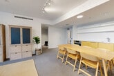 Located in Vancouver's West End at V6G 1W1, this contemporary amenity room offers a well-appointed, single-floor open-plan layout designed for communal gathering and casual use. The space is finished with neutral grey carpeting throughout and crisp white walls, creating a clean and inviting atmosphere.

The room is divided into two functional zones. The dining area features a long natural wood table paired with matching honey-toned folding chairs, comfortably seating multiple guests. Adjacent to it sits a compact kitchenette with warm yellow cabinetry, a built-in stainless steel oven, and a sink — ideal for catering light refreshments. Built-in open shelving above provides additional storage and display space.

The lounge area is anchored by a cream sofa and a sleek black marble coffee table, positioned to take full advantage of the expansive floor-to-ceiling windows. These large glazed panels flood the space with natural light and offer pleasant views of a landscaped courtyard and the surrounding neighbourhood streetscape. A large monochromatic landscape painting adorns one wall, adding an artistic focal point.

Recessed pot lights and adjustable track lighting provide versatile illumination throughout. A light wood storage cabinet with blue-panelled doors adds both function and a subtle pop of colour. The overall ambiance is modern, bright, and welcoming — well-suited for residents to meet, work, or entertain.