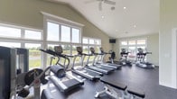 Located at 3381 Blue Heron Dr in Hilliard, Ohio, this single-floor fitness center is designed to offer a bright, welcoming environment for exercise and wellness. The space is defined by its striking vaulted ceiling, which lends a sense of openness and volume to the interior. A ceiling fan is centrally mounted overhead, providing comfortable air circulation throughout workouts. Recessed lighting complements the abundant natural light that pours in through a generous array of large white-trimmed windows lining the walls, offering pleasant views of the surrounding green outdoor landscape. The flooring features durable, dark rubber matting appropriate for a high-use fitness environment. The facility is well-equipped with a row of treadmills and elliptical machines positioned to take advantage of the outdoor views, alongside a stationary bike, stair climber, and free weight equipment including a weight bench. A wall-mounted television is also present for entertainment during workouts. The sage green walls contribute to a calm, clean aesthetic that complements the bright, airy atmosphere created by the vaulted ceiling and expansive windows. Overall, the fitness center provides a functional yet inviting space that encourages an active lifestyle.