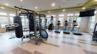 This fitness center is designed to provide a comprehensive workout environment within a bright, open single-floor layout. The space is defined by its striking interior design, featuring bold chartreuse green accent beams contrasted against crisp white walls, creating an energetic and motivating atmosphere. A multi-colored checkerboard-patterned floor in neutral tones with green accents ties the design together throughout.

Large windows line multiple walls, flooding the space with abundant natural light and offering views of the surrounding outdoor environment. Ceiling fans and recessed lighting provide additional comfort and illumination.

The cardio section is well-appointed with multiple treadmills, ellipticals, and stationary bikes, each equipped with individual display screens. A stair climber rounds out the cardiovascular offerings. The strength training area features TKO-branded cable and multi-functional machines, a power rack, kettlebells, a BOSU ball, a mini trampoline, and a full dumbbell rack. A weight bench provides additional versatility for free weight exercises.

Full-length mirrors along one wall expand the sense of space and allow users to monitor their form. The facility also includes a dedicated stretching and open floor area, as well as a small storage unit near the entrance. The space is accessible via multiple doors and maintains a clean, organized layout throughout.