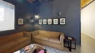The Resident Lounge is a thoughtfully designed communal space that blends industrial aesthetics with comfortable, inviting furnishings. The room features soaring ceilings with exposed wooden beams and visible structural elements, lending an urban, loft-like character to the space. Overhead, a combination of pendant lights with aged metal dome shades and wall-mounted sconces cast a warm, intimate glow throughout.

Deep slate-blue walls set a sophisticated, moody tone and serve as an elegant backdrop for a curated gallery of framed vintage-style artwork arranged in a neat row. The seating area centers around generously sized, tufted tan leather sofas arranged in an L-shaped configuration, offering ample room for socializing or relaxing. A patterned geometric area rug in muted tones anchors the seating arrangement, adding texture underfoot, while a rustic wooden coffee table provides a functional focal point.

Natural light filters in through a framed window, providing a soft counterbalance to the atmospheric artificial lighting. Polished concrete flooring extends throughout, reinforcing the industrial character of the space. The overall ambiance is sophisticated yet comfortable, creating an ideal setting for residents to unwind or gather.
