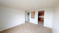 This single-floor residence offers a straightforward and functional layout designed for comfortable everyday living. The main living area is finished with wall-to-wall beige carpeting, creating a warm and quiet environment. Crisp white walls and a smooth ceiling keep the space feeling bright and open throughout.

A distinctive architectural pass-through window connects the living area directly to the kitchen, promoting an open feel while maintaining a defined separation between the two spaces. The kitchen is outfitted with warm-toned wood cabinetry featuring both upper and lower storage units, complemented by tile flooring that adds durability and easy maintenance to the cooking area. A ceiling fan is visible in the kitchen, providing additional air circulation.

Adjacent to the main living space, a hallway leads to a generously sized closet with bifold doors, offering ample storage for the unit. The flooring transitions from carpet to tile in this connecting corridor, reinforcing the practical zoning of the home.

Overall, the space presents a clean, neutral palette that serves as a versatile backdrop, making it easy to personalize. The combination of carpeted living spaces, tiled functional areas, and connected kitchen layout gives this residence a classic and livable character suited to a variety of lifestyles.