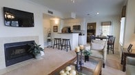 This single-floor residence offers a thoughtfully designed open-concept layout that promotes a natural flow between living, dining, and kitchen areas. The spacious living room is anchored by a fireplace and features plush carpeting, crown molding, and a wall-mounted television, creating a comfortable and polished atmosphere. A glass-top coffee table and neutral-toned furnishings add an understated elegance to the space. The kitchen is positioned centrally and is equipped with white cabinetry, stainless steel appliances including a microwave and range, and a breakfast bar with bar stool seating — ideal for casual dining or entertaining. Recessed lighting and track lighting illuminate the space efficiently throughout. Crown molding runs consistently across the ceiling, reinforcing the refined character of the interior. A glimpse into the adjoining bedroom reveals additional living space with natural light filtering through a window with tailored treatments. The overall ambiance is warm and welcoming, with a cohesive neutral color palette, carpeted floors, and classic architectural details that lend the home a timeless residential appeal.