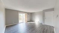 This single-floor unit presents a clean, contemporary aesthetic throughout, unified by light grey vinyl plank flooring and a neutral grey-and-white color palette. The spacious living area is a standout feature, offering a vertical shiplap-paneled accent wall, baseboard heating, a wall-mounted air conditioning unit, and a sliding glass door that opens onto a private wooden deck with outdoor views of mature trees—bringing in generous natural light.

The galley-style kitchen is efficiently designed with white cabinetry, granite-look countertops, a stainless steel sink, and a full-size refrigerator. A passthrough area connects the kitchen to the broader living space, enhancing the open feel of the floor plan.

The unit includes multiple bedrooms, each finished with the same grey vinyl flooring, neutral walls, and baseboard heating. One bedroom features a built-in closet with shelving, while another benefits from a bright window that admits ample natural light. A dedicated entry area provides a formal point of arrival. The bathroom is outfitted with a full tub-shower combo, a toilet, and a granite-topped vanity with white cabinetry, maintaining the unit's consistent modern finish throughout.