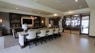 This single-floor space exudes a polished, contemporary elegance through its thoughtful design and high-end finishes. The centerpiece of the space is a generously sized bar counter finished in light granite, lined with a row of cushioned, cream-upholstered bar chairs featuring dark wood frames and nailhead trim detailing — a refined touch that anchors the lounge aesthetic.

Behind the bar, dark cabinetry provides a dramatic backdrop, complemented by ambient backlighting, decorative glassware display, and a large wall-mounted flat-screen television. A sculptural, globe-cluster chandelier hangs overhead, adding an artistic focal point that balances the warmth of wall sconces nearby.

The flooring throughout is composed of large-format light tile, contributing to an open, airy feel that contrasts beautifully with the darker wall treatments. Recessed lighting and tray ceiling details enhance the architectural depth of the space.

To the right, floor-to-ceiling glass partitions with dark metal framing open into an adjacent area, allowing natural light to filter through while maintaining a sense of defined zones. The overall ambiance is sophisticated and welcoming, suited for social or hospitality-oriented use.