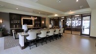This single-floor space exudes a polished, contemporary elegance through its thoughtful design and high-end finishes. The centerpiece of the space is a generously sized bar counter finished in light granite, lined with a row of cushioned, cream-upholstered bar chairs featuring dark wood frames and nailhead trim detailing — a refined touch that anchors the lounge aesthetic.

Behind the bar, dark cabinetry provides a dramatic backdrop, complemented by ambient backlighting, decorative glassware display, and a large wall-mounted flat-screen television. A sculptural, globe-cluster chandelier hangs overhead, adding an artistic focal point that balances the warmth of wall sconces nearby.

The flooring throughout is composed of large-format light tile, contributing to an open, airy feel that contrasts beautifully with the darker wall treatments. Recessed lighting and tray ceiling details enhance the architectural depth of the space.

To the right, floor-to-ceiling glass partitions with dark metal framing open into an adjacent area, allowing natural light to filter through while maintaining a sense of defined zones. The overall ambiance is sophisticated and welcoming, suited for social or hospitality-oriented use.