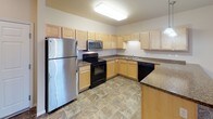 This single-floor residence offers a well-designed, open-concept layout that seamlessly connects the kitchen and living area. The kitchen is outfitted with light maple cabinetry, granite-style countertops, stainless steel and black appliances including a refrigerator, electric range, microwave, and dishwasher, and a tile floor in a warm earth-tone pattern. A peninsula counter with pendant lighting above serves as both a workspace and a natural divider between the kitchen and the carpeted living area. A built-in desk nook with under-cabinet lighting adds a functional workspace near the kitchen. The living area benefits from natural light through a sliding glass door that opens to the exterior. Bedrooms are carpeted and generously sized, each featuring windows that let in natural light and closets — including at least one walk-in closet with wire shelving systems. The bathroom features a full tub-and-shower combination, a vanity with granite-style countertops and maple cabinetry, and tile flooring consistent with the rest of the home. A dedicated in-unit laundry area accommodates a full-size washer and dryer side by side, complete with overhead wire shelving. Baseboard heating is present throughout, and the neutral warm-toned walls contribute to a clean, move-in-ready aesthetic.