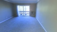 This single-floor space offers a straightforward and functional layout designed for comfortable everyday living. The main living area is finished with wall-to-wall carpeting, providing a soft and cohesive look throughout. A prominent sliding glass door serves as the focal point of the room, allowing an abundance of natural light to flood the interior while offering direct access to an outdoor deck. The deck, framed by a wooden railing, overlooks a lush, tree-lined outdoor setting, creating a pleasant connection between the interior and the surrounding environment. Sheer curtains hang from a rod above the sliding door, offering privacy and light diffusion as needed. The walls are painted in a neutral, earthy tone complemented by clean white crown molding and wide baseboards, lending the space a classic and tidy appearance. A wall-mounted air conditioning unit is conveniently positioned near the sliding door, ensuring year-round climate control. The overall ambiance of the space is clean, bright, and ready for personalization, making it a versatile and practical living environment.