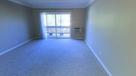 This single-floor space offers a straightforward and functional layout designed for comfortable everyday living. The main living area is finished with wall-to-wall carpeting, providing a soft and cohesive look throughout. A prominent sliding glass door serves as the focal point of the room, allowing an abundance of natural light to flood the interior while offering direct access to an outdoor deck. The deck, framed by a wooden railing, overlooks a lush, tree-lined outdoor setting, creating a pleasant connection between the interior and the surrounding environment. Sheer curtains hang from a rod above the sliding door, offering privacy and light diffusion as needed. The walls are painted in a neutral, earthy tone complemented by clean white crown molding and wide baseboards, lending the space a classic and tidy appearance. A wall-mounted air conditioning unit is conveniently positioned near the sliding door, ensuring year-round climate control. The overall ambiance of the space is clean, bright, and ready for personalization, making it a versatile and practical living environment.