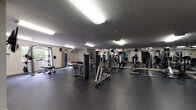 The fitness center at Stadium Tower is a comprehensively outfitted facility designed to support a full range of workout routines. The space is anchored by durable rubber flooring throughout, providing a safe and functional surface for all types of exercise. The main workout area is generously sized, featuring an extensive array of strength-training equipment including cable machines, multi-function weight stations, a barbell rack, and a free weights section. A full wall of mirrors lines one side of the space, amplifying the sense of openness and allowing users to monitor their form during workouts. The cardio zone is equipped with treadmills, elliptical trainers, and stationary bikes, several of which are fitted with individual screens for entertainment during workouts. Flat-screen televisions are mounted throughout the facility, and a dedicated functional fitness corner houses stability balls, resistance bands, and a wall-mounted barbell station. Motivational wall decals, including phrases like "Beast Mode On" and "The body achieves what the mind believes," add an energetic atmosphere to the space. Overhead fluorescent lighting ensures the entire facility is well-lit. The fitness center is accessible directly from the parking structure, adding convenience for residents and users of Stadium Tower.