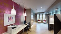 This contemporary single-floor office space combines bold design elements with practical functionality. The workspace is immediately distinguished by a striking textured magenta accent wall that runs along the workstation area, creating a vibrant focal point throughout the space. Suspended pendant lights with decorative white fixtures illuminate a row of built-in desks, providing an efficient and stylish work area. Glass partitions divide the workspace, maintaining an open and airy feel while offering defined zones for different activities.

A bright lounge area features large windows that invite generous natural light, furnished with a pair of elegant tufted white armchairs accented with deep red throw pillows, creating an inviting reception or relaxation space. Light wood flooring extends throughout this area, enhancing the sense of openness.

Two conference rooms serve collaborative needs. One room is equipped with a long white table surrounded by bold magenta chairs, complemented by a wall-mounted flat-screen television set against the signature magenta textured wall. The second conference room features a white table with matching pink chairs and a creative arrangement of hexagonal whiteboard panels mounted on the wall, serving both as a functional writing surface and a distinctive design feature. Recessed lighting and carpet flooring complete these professional meeting spaces.