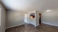 This single-story, one-bedroom, one-bathroom unit in Houston, TX (77090) offers a practical and well-appointed layout designed for comfortable living. The main living area is finished with rich, dark hardwood-style flooring and neutral gray walls, creating a clean, modern aesthetic throughout. A pass-through window with accent lighting connects the living space to the kitchen, providing an open feel between the two areas. The galley-style kitchen is efficiently designed, featuring dark cabinetry, light stone-patterned countertops, a stainless steel refrigerator, and an electric range, all beneath a textured ceiling with built-in ventilation. The bedroom is carpeted and includes a ceiling fan with a window dressed in blinds that allows natural light to filter in. A generously sized walk-in closet with wrap-around shelving and hanging rods provides ample storage. The bathroom includes a full tub-and-shower combination with tiled surround. White trim and baseboards run consistently throughout the unit, complementing the neutral color palette. The overall ambiance is clean, functional, and move-in ready, suited for single occupants or couples seeking a manageable, low-maintenance living space in Houston.