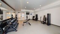 Located at 8585 La Mesa Blvd in La Mesa, San Diego, this well-appointed fitness center offers a comprehensive workout facility on a single level. The space is thoughtfully divided into distinct exercise zones, providing a well-rounded experience for users of all fitness levels.

The cardio area is bathed in natural light through large sliding glass windows and doors that open directly to an outdoor courtyard, creating a bright and airy atmosphere. This zone is equipped with a treadmill and an elliptical trainer, with additional cardio options including a stationary cycle nearby.

The strength training section features a full rack of dumbbells displayed on a mirrored wall, an adjustable weight bench, and multiple selectorized machines including a leg press and additional cable-based resistance units. A wall-mounted flat-screen television and a built-in water fountain station add convenience to this area, which is accessible via glass-paneled French doors.

The space is finished with durable, light-toned flooring throughout, recessed ceiling lighting, and a clean, neutral color palette that contributes to an open and motivating environment. Glass-panel entry doors provide transparency between zones, and direct access to the outdoor area reinforces the connection to the surrounding community. The overall design is modern, functional, and well-suited for daily fitness use.
