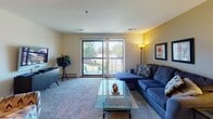 Located at 1600 Pointe Dr in Valparaiso, Indiana, this single-floor residence offers a well-organized layout designed for comfortable everyday living. The living room is a generous, carpet-floored space anchored by a large sectional sofa and a glass-top coffee table. A bold accent wall adds a warm, inviting character, while a sliding glass door opens to an outdoor patio, filling the room with natural light and providing pleasant outdoor views. The kitchen is efficiently designed with rich wood cabinetry, white tile backsplash, light tile flooring, and stainless steel appliances including a refrigerator, dishwasher, and range. Adjacent to the kitchen, the dining area features a round glass-top table with a ceiling fan overhead, creating a cozy setting for meals. The primary bedroom is a calm retreat with carpet flooring, a window dressed in warm drape curtains, and a spacious closet. A second bedroom mirrors this comfort with its own closet and natural light. Two bathrooms serve the residence: one with a walk-in shower and tile flooring, and another with a tub-shower combination, vanity sink, and ample mirror. Throughout, neutral tones and practical finishes create a cohesive, livable atmosphere well-suited for a variety of lifestyles.