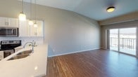 This single-floor unit offers a well-designed open-concept layout anchored by a contemporary kitchen and expansive living area. The kitchen is equipped with stainless steel appliances, including a refrigerator, electric range, and over-the-range microwave, complemented by white shaker-style cabinetry, a subway tile backsplash, and a peninsula island with a light stone countertop and pendant lighting above. The soaring ceilings in the main living area lend a spacious, airy quality to the space, with warm-toned hardwood flooring extending throughout.

A full bathroom features a granite-topped vanity with white cabinetry, a tub-shower combination, and hardwood flooring. A separate half bath is accessible from the main living corridor. The bedroom is carpeted and receives natural light through a window with exterior views. A walk-in closet adjacent to the bathroom provides wire shelving on both sides for ample storage.

The unit also includes a balcony accessible from the living area, visible in the overhead view. Neutral warm-gray walls throughout create a cohesive, modern aesthetic, while white trim and panel doors add a clean, polished finish to every room.