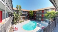 This multi-unit residential complex is thoughtfully organized around a central courtyard that serves as the heart of the community. The standout feature of the courtyard is a generously sized, kidney-shaped swimming pool with clear turquoise water, surrounded by a clean pool deck furnished with lounge chairs and an outdoor grill — ideal for relaxation and socializing. Mature trees and tropical plantings, including banana palms and ornamental shrubs, add greenery and shade throughout the courtyard, contributing to a warm, resort-like atmosphere.

The building spans multiple stories and is arranged in a U-shaped configuration that wraps around the courtyard, maximizing views and connectivity. Each unit is accessed via open-air exterior corridors that run the length of the building, offering direct sightlines to the pool below. The corridors are finished in a textured stucco exterior with large windows allowing natural light into each unit. A recurring design element throughout the property is the use of bold red accent doors, which provide a striking visual contrast against the neutral grey and tan tones of the building's facade. Unit numbers are clearly marked on each door. The overall aesthetic is clean, functional, and visually cohesive, reflecting a well-maintained residential community.