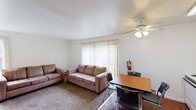This single-floor unit offers a practical and straightforward layout designed for comfortable everyday living. The living room serves as the central gathering space, finished with wall-to-wall carpet and outfitted with two sofas and a ceiling fan with integrated lighting. A large sliding glass door flanked by vertical blinds fills the room with natural light and provides views of surrounding greenery. A dining area with a wood-toned table and chairs is positioned nearby, creating a seamless transition between the living and eating spaces.

The kitchen features white cabinetry extending to the ceiling for ample storage, white appliances including a range, dishwasher, and refrigerator, a single-basin sink, and light countertops. Wood-style flooring and a ceiling fan complete the functional space.

The bedroom is a generously sized, carpeted room with clean white walls, a baseboard heater, and an adjacent walk-in closet equipped with dual hanging rods and shelving on multiple levels, providing substantial storage capacity.

The bathroom includes a dual-sink vanity with a wide mirror above, tile flooring, and a separate tub/shower combination enclosed with mosaic-tile surround walls. The overall design throughout the unit is clean and neutral.