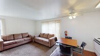 This single-floor unit offers a practical and straightforward layout designed for comfortable everyday living. The living room serves as the central gathering space, finished with wall-to-wall carpet and outfitted with two sofas and a ceiling fan with integrated lighting. A large sliding glass door flanked by vertical blinds fills the room with natural light and provides views of surrounding greenery. A dining area with a wood-toned table and chairs is positioned nearby, creating a seamless transition between the living and eating spaces.

The kitchen features white cabinetry extending to the ceiling for ample storage, white appliances including a range, dishwasher, and refrigerator, a single-basin sink, and light countertops. Wood-style flooring and a ceiling fan complete the functional space.

The bedroom is a generously sized, carpeted room with clean white walls, a baseboard heater, and an adjacent walk-in closet equipped with dual hanging rods and shelving on multiple levels, providing substantial storage capacity.

The bathroom includes a dual-sink vanity with a wide mirror above, tile flooring, and a separate tub/shower combination enclosed with mosaic-tile surround walls. The overall design throughout the unit is clean and neutral.