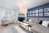 This thoughtfully designed single-floor residence offers a cohesive and modern interior highlighted by light wood flooring that flows seamlessly through every room. The living room serves as the heart of the home, featuring a fireplace framed by a wood mantle shelf, a bold navy accent wall adorned with a gallery of framed artwork, and natural light streaming in through windows dressed with patterned curtains and wood blinds. A cozy reading nook with a lounge chair adds functional charm to the space.

The kitchen is outfitted with white shaker-style cabinetry, stainless steel appliances including a refrigerator, range, and microwave, as well as a single-basin sink with a modern faucet and ample counter space. The adjacent dining area comfortably accommodates a four-person table beneath a ceiling fan with light fixture, with exterior door access and gold-framed mirror accents on the wall.

Both bedrooms feature accent walls in rich blue tones, providing a restful and stylish atmosphere, each with natural light from windows with blinds and curtains. A hallway lined with built-in closet storage connects the rooms efficiently. The full bathroom includes a shower-tub combination and is accented with framed wall art, completing this well-organized and visually cohesive residence.