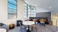 Located at 1130 E 3rd Ave in Oakland, California, this contemporary two-floor lounge space offers a thoughtfully designed environment spread across two distinct levels. The lower floor serves as an entry and transition area, featuring plush upholstered seating, a sleek staircase with a polished stainless steel cable railing, and a bold navy accent wall that adds a striking visual contrast to the crisp white surroundings. Textured, multi-toned carpet flooring runs throughout, adding warmth and acoustic comfort underfoot. A glass door on this level provides direct access to the outdoors, while a glimpse into an adjacent fitness area hints at a broader amenity-rich environment. The upper floor serves as the primary gathering space, characterized by soaring vaulted ceilings that amplify the sense of openness. Floor-to-ceiling windows flood the room with natural light and frame views of neighboring greenery and the surrounding Oakland streetscape. A geometric patterned accent wall in muted gray and gold tones anchors one side of the room, complementing the modern furnishings that include round marble-top tables, mixed dining chairs, and a built-in black leather banquette. Recessed lighting and strategically placed overhead fixtures ensure a well-lit atmosphere throughout. The overall ambiance is sleek, inviting, and well-suited for communal gathering or relaxed socializing.