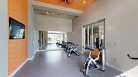 The Spin Room is a dedicated indoor cycling studio designed to deliver an energizing and focused workout experience. The space features a row of stationary spin bikes arranged to face a full wall of mirrors, which visually expand the room while allowing cyclists to monitor their form during sessions. Soaring ceilings with a distinctive slatted wood ceiling panel in warm orange tones add architectural character and lend a modern, dynamic ambiance to the space.

Large windows along one wall flood the studio with natural light and provide views of outdoor greenery and the surrounding area, contributing to an open and motivating atmosphere. The durable gray flooring is well-suited for high-intensity workouts, providing a clean and low-maintenance surface throughout.

An accent wall clad in orange horizontal slat paneling serves as a focal point, featuring a wall-mounted flat-screen television for instructor-led virtual classes or entertainment during rides. TRX suspension training straps and wall-mounted equipment hooks offer additional cross-training options beyond cycling. Glass-paneled doors connect the Spin Room to an adjacent fitness area, ensuring a seamless transition between workout spaces. Recessed lighting complements the natural light for a well-illuminated environment at any time of day.