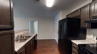This single-floor residence offers a functional and cohesive layout designed for comfortable everyday living. The kitchen is a standout space, featuring dark espresso cabinetry, granite-look countertops, and a full suite of black appliances including a side-by-side refrigerator, electric range with overhead microwave, and dishwasher. A breakfast bar connects the kitchen to the open living and dining area, which is finished with rich, dark hardwood-style flooring and illuminated by an elegant chandelier. Natural light filters in through a sliding door, enhancing the airy feel of the common areas. Two bathrooms mirror the kitchen's design aesthetic, each with dark wood vanities, granite-look countertops, and large mirrors. One bathroom includes a walk-in shower with wood-style flooring, while the second provides convenient access from the main living areas. The bedrooms are finished with plush neutral carpet and feature closets with wire shelving for practical storage. A dedicated laundry area is equipped with washer and dryer hookups, adding to the unit's overall convenience. Neutral wall tones and consistent finishes throughout create a clean, move-in ready environment.