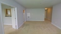 This single-floor residential space offers a straightforward, functional layout designed with simplicity and comfort in mind. The main living area is characterized by soft, wall-to-wall beige carpeting that adds warmth underfoot, while the light gray walls create a neutral and versatile backdrop throughout. The open floor plan allows for a seamless flow between areas, providing flexibility in how the space can be arranged and utilized.

Recessed vents and ceiling-mounted light fixtures are integrated throughout, ensuring consistent climate control and ambient lighting. The smooth white baseboards and clean wall finishes reflect a well-maintained interior. An open doorway leads to a hallway, connecting additional areas of the space and contributing to its overall sense of openness.

Visible from the main area, a secondary room or utility space is accessible through an adjacent doorway, partially revealing what appears to be a refrigerator, suggesting a kitchen or storage area nearby. A wall panel — likely a thermostat or utility access point — is also present, adding to the practical nature of the layout.

Overall, the space presents a clean, move-in ready environment with a calm, neutral aesthetic that offers a solid foundation for personalization and furnishing.