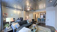 This single-floor studio showcases a distinctive industrial-chic aesthetic, anchored by a standout metallic ceiling with exposed ductwork that adds raw architectural character to the space. A ceiling fan with integrated lighting serves as a functional centerpiece overhead.

The open-plan layout is thoughtfully divided into distinct zones without the use of permanent walls. A sheer curtain panel elegantly separates the sleeping area from the living space, offering a soft, airy sense of privacy. The sleeping zone features a full-size bed dressed in vibrant coral and pink bedding, flanked by a curated gallery wall that adds personality and warmth. A dedicated workspace with a desk and chair is tucked neatly into one corner.

The living area hosts a spacious grey sectional sofa paired with a glass-top coffee table and a patterned area rug, creating a comfortable and inviting lounge space. A classic Eames-style lounge chair and ottoman add a touch of mid-century modern sophistication.

Toward the back, a kitchen with a breakfast bar and pendant lighting provides a polished transition into the culinary space, complete with bar-height seating. Dark hardwood-style flooring runs throughout, tying all the zones together cohesively. The overall ambiance balances urban industrial elements with warm, personalized décor.
