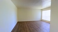 This single-floor space presents a straightforward, open layout well-suited for a variety of uses. The main living area is defined by warm, dark hardwood-style plank flooring that runs throughout, providing a grounded, cohesive aesthetic that contrasts cleanly with the light, neutral-toned walls and ceiling. The walls are finished in a soft off-white, creating a bright and airy atmosphere that makes the room feel open and versatile. A window positioned along one side of the room admits natural light, softened by vertical blinds that allow for adjustable privacy and light control. The overall design is minimal and functional, with clean baseboards framing the perimeter of the room. The absence of ornamental detailing keeps the space feeling modern and adaptable. The room provides ample open floor area, offering flexibility for various furniture arrangements and layouts. Overall, the space maintains a clean, neutral palette that serves as a practical and welcoming foundation for personalization.