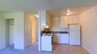 This single-floor space offers a practical and straightforward layout designed for everyday comfort. The kitchen serves as a central feature, outfitted with white upper and lower cabinetry that provides generous storage. A full suite of appliances is included — an electric range with coil burners, a dishwasher, and a full-size refrigerator — all in a coordinating white finish. Dark countertops offer a subtle contrast against the light cabinetry, and an overhead range hood is positioned above the stovetop. A double-basin sink sits beneath additional upper cabinets, completing the functional workspace. The kitchen flows openly toward the living area, where light-toned carpeting adds a soft underfoot feel. Tile flooring in the kitchen area provides a durable surface suited for daily use. A textured ceiling runs throughout, and flush-mount light fixtures offer consistent overhead illumination. A hallway visible from the kitchen leads to additional rooms beyond, suggesting a well-organized floor plan. The overall aesthetic is clean and neutral, with a simple, move-in-ready character suited to a wide range of occupants.