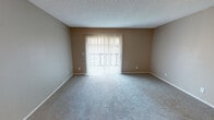 This single-floor unit is designed with a practical, open layout that connects the main living area to the kitchen. The living room is generously sized, finished with plush neutral-toned carpet and warm beige walls. A large sliding glass door fitted with vertical blinds fills the space with natural light while providing outdoor access. A textured ceiling adds a subtle architectural detail consistent throughout the unit.

The kitchen runs along a compact galley-style corridor, featuring white cabinetry, dark countertops, and a blue tile backsplash. Black appliances — including a refrigerator, electric range, and dishwasher — contrast cleanly against the white cabinets. Wood-look flooring runs through the kitchen, adding warmth and durability to the space.

The bedrooms are carpeted and finished in the same neutral palette, each featuring windows with blinds that allow natural light to filter in. The rooms offer ample wall space suitable for various furniture arrangements.

The bathroom is well-appointed with a white vanity cabinet, dark countertop, large mirror, and a multi-bulb vanity light fixture. A full tub-shower combination with white tile surround and wood-look flooring completes the bathroom's clean, functional aesthetic.