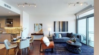 This single-floor residence features an open-concept layout that elegantly combines the living and dining areas into one cohesive, functional space. The living area is anchored by a blue-grey sofa accented with teal throw pillows, complemented by a rust-orange accent chair draped with a cozy faux-fur throw. A round, dark-toned coffee table with a sculptural base serves as a central focal point, set atop a rich navy blue area rug that adds depth and contrast to the rich hardwood flooring beneath. A large abstract painting in blues, golds, and greys decorates the wall, enhancing the contemporary aesthetic. The dining area nearby features a round table with cream upholstered chairs, alongside a mid-century modern sideboard topped with decorative accents and a framed geometric mirror. Track lighting runs across the white ceiling, providing versatile, well-distributed illumination throughout. Large windows allow natural light to filter into the space, brightening the crisp white walls and creating an airy, welcoming ambiance throughout.