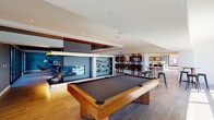 This single-floor space is thoughtfully designed with a contemporary aesthetic, offering a diverse range of amenity areas that flow seamlessly into one another. The open layout connects several distinct zones, each with its own character while maintaining a cohesive modern design language throughout.

The game room anchors the space with a full-size billiard table set atop wood-look tile flooring. An illuminated built-in shelving unit with a sleek fireplace serves as a central architectural feature, finished in slate-toned tile and framed by warm wood accents. Dark wood slatted ceilings define the adjacent lounge area, which features a vibrant teal accent wall, large-format tile flooring, and floor-to-ceiling sliding glass doors that open to an outdoor terrace, flooding the space with natural light.

The kitchen is outfitted with stainless steel appliances, rich walnut cabinetry with frosted glass uppers, a herringbone tile backsplash, and a striking island combining a marble base with a live-edge wood countertop. Statement gold pendant lighting adds warmth overhead. A separate windowed nook with a high-top table offers panoramic outdoor views.

A dedicated golf simulator room completes the space, featuring a mounted projector, a projection screen, and a putting mat — ideal for recreational use. Throughout, the design balances bold color choices with clean lines and high-quality materials.