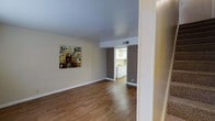 This two-floor space offers a clean, functional layout with a welcoming aesthetic. The main living area is defined by warm hardwood-style flooring that runs throughout, complementing the neutral beige walls and light grey ceiling. A colorful decorative piece on the wall adds a subtle artistic touch to the otherwise understated palette. The space feels open and uncluttered, with smooth transitions between areas.

Visible from the living area, the kitchen is accessible through a wide opening framed by contrasting dark grey accent walls, creating a natural visual division between the two spaces. The kitchen features white cabinetry and white appliances, contributing to a bright, tidy appearance. Overhead lighting within the kitchen ensures the space remains well-lit.

Recessed ceiling vents are present throughout, supporting comfortable climate control. White baseboards provide clean contrast against the flooring and walls. The overall ambiance is straightforward and practical, with a neutral color scheme that offers versatility for a variety of interior styles. The two-floor configuration further adds to the functional depth of the overall space.