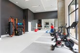 Located on Halcyon Way in Alpharetta, Georgia, this single-floor fitness center offers a comprehensive and thoughtfully designed workout facility. The main gym area features soaring ceilings with an eye-catching coffered metal grid framework, exposed concrete columns, and floor-to-ceiling windows that flood the space with natural light and provide views of the surrounding outdoor terrace. The gym is well-equipped with a full row of cardio machines including treadmills and ellipticals, along with cable machines, adjustable benches, and a full dumbbell rack. A dedicated free weights and functional training zone sits adjacent to a service counter and kettlebell storage. A separate studio room is finished with sleek dark accent walls, full-length mirrored walls, a ballet barre, stability balls, and BOSU trainers — ideal for group fitness or individual training sessions. The spin studio features multiple stationary bikes with digital displays, positioned in front of large windows overlooking an outdoor balcony. Throughout the facility, light-toned flooring with contrasting stripe detailing provides a clean, modern aesthetic. The overall design balances industrial and contemporary elements to create an energizing and functional fitness environment.