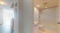 This well-designed single-floor unit offers a thoughtfully laid-out interior with a clean, contemporary aesthetic throughout. The open-concept living area is anchored by warm hardwood flooring and illuminated by track lighting overhead, with floor-to-ceiling windows dressed in vertical blinds that invite abundant natural light and provide views of the surrounding cityscape. The living space flows seamlessly into a modern kitchen, which features dark granite countertops, warm wood cabinetry with glass-front upper cabinets, and stainless steel appliances including a Bosch microwave, electric cooktop, and oven. Large-format tile flooring completes the kitchen's polished look. The bedrooms are finished with plush carpeting and benefit from the same expansive windows, ensuring natural light and outdoor views. The unit includes two bathrooms: a primary bathroom featuring a marble-topped double vanity, large-format white tile flooring, a glass-enclosed shower, and a soaking tub; and a secondary bathroom with white tile flooring, a combined shower and bathtub with large tile surround, and clean white fixtures throughout. The overall ambiance is bright, minimal, and modern, offering a versatile canvas ready for personalization.