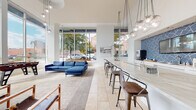 This thoughtfully designed single-floor space blends contemporary aesthetics with functional amenities, creating an inviting environment suited for both relaxation and social interaction. The layout flows seamlessly from a formal lobby entrance to open living and entertaining areas.

The lobby sets an immediate tone of sophistication, featuring herringbone-patterned travertine flooring, soaring ceilings, and sculptural ring pendant lighting. A clean white reception desk anchors the entry alongside curated wall art, establishing a polished first impression.

The central lounge area is expansive and light-filled, framed by floor-to-ceiling windows that offer views of surrounding streetscapes and mature trees. A bold blue sectional sofa anchors the seating zone atop a neutral area rug, complemented by mid-century inspired wooden chairs and a wall-mounted television. A foosball table adds a recreational element near the glazed perimeter.

A long, curved bar counter with industrial-style metal and wood barstools extends along one side of the lounge. Above, clusters of globe pendant lights in varying heights create a dramatic visual feature. Adjacent to the bar, a fully equipped kitchenette showcases a striking blue geometric tile backsplash, white cabinetry, marble-look countertops, a stainless double sink, and built-in appliances including a coffee station and oven.