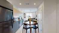 This contemporary single-floor residence showcases a thoughtfully designed open-concept layout that seamlessly connects the kitchen, dining, and living areas. The kitchen is a standout feature, equipped with full-height light wood upper cabinetry, matte grey lower cabinets, a stone-look backsplash with integrated under-cabinet lighting, and a complete suite of stainless steel appliances including a double oven, built-in microwave, dishwasher, and refrigerator. A round dining table anchors the transition between the kitchen and the bright living room, which features a ceiling fan, recessed lighting, and large windows offering urban views. Light-toned hardwood flooring flows consistently throughout the space, enhancing its cohesive, airy feel. The primary bedroom is characterized by warm taupe accent walls, a ceiling fan, and a dedicated closet with mirrored sliding doors and built-in shelving. The second bedroom features a bold deep green accent wall, its own closet, and windows with natural light and outdoor views. Both bathrooms are finished to a high standard, featuring large-format grey stone-look tile, glass-enclosed walk-in showers with built-in benches, multi-function matte black shower panels, floating vanities with white countertops, and backlit mirrors.