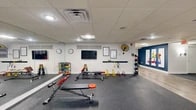 This gym in Port Charlotte, Florida, offers a thoughtfully organized fitness facility spread across a single floor. The layout is anchored by a primary workout area featuring rubber flooring, large wall-to-wall mirrors, and a dropped tile ceiling with recessed and flush-mounted lighting that ensures a bright, energizing environment. This zone is outfitted with a comprehensive range of equipment, including adjustable benches, a dumbbell rack with color-coded weights, kettlebells, medicine balls, resistance bands, agility hurdles, and a wall-mounted flat-screen TV for workout guidance. A wall-mounted industrial fan provides added airflow and comfort during training sessions.

The space transitions into a wide, open hallway finished with light wood-look flooring and bold navy blue accent walls adorned with colorful fitness-themed artwork. This corridor serves a dual purpose, functioning as both a circulation path and an extension of the fitness area, with yoga mat storage racks, stability balls, and a water fountain conveniently accessible along the walls. The hallway connects to a cardio zone featuring treadmills and elliptical machines positioned near windows that invite natural light. Colorful seating accents and framed informational signage add personality and practicality throughout the space.
