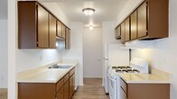 This single-floor residence offers a functional and straightforward layout across its main living areas. The galley-style kitchen is a standout feature, lined on both sides with warm wood-toned cabinetry and light countertops that provide ample storage and prep space. It is equipped with white appliances including a gas range and refrigerator, a single-basin sink, and a dishwasher, all set atop durable wood-look flooring that runs the length of the kitchen. Overhead flush-mount lighting keeps the space bright and practical.

The living area is generously proportioned, featuring wall-to-wall neutral carpeting and crisp white walls that create a clean, open feel. Large vertical blinds cover a full-width window, allowing for adjustable natural light throughout the day. A ceiling fan with a light fixture adds comfort and function to the space.

The bedroom is a well-sized, carpeted room with white walls and a dark wood interior door, offering a quiet and private retreat. Adjacent to the bedroom is a full bathroom appointed with white tile flooring, a single vanity with wood cabinetry and a mirrored medicine cabinet, a toilet, and a shower-tub combination. A lighted vanity bar provides ample illumination. The 3D dollhouse view also suggests the presence of a closet or storage area within the unit's layout.