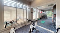 This well-appointed fitness studio is designed to support a comprehensive workout experience within a single, thoughtfully organized space. The room is finished with blue textured carpeting underfoot and neutral, subtly textured wall coverings that lend a clean, polished aesthetic throughout.

A dedicated cycling area anchors one end of the studio, equipped with True brand stationary bikes positioned to face a large wall-mounted flat-screen television, ideal for guided rides or entertainment during workouts. A wall-mounted control panel is conveniently located nearby.

Large floor-to-ceiling glass panel windows and a glass door connect the fitness studio to an adjacent lounge or common area, allowing natural light to flow generously into the space and creating a bright, open atmosphere. Clerestory windows above further enhance the sense of airiness.

Full-length wall mirrors on one side amplify the perceived depth of the room while providing functional visibility during exercise. Additional equipment is visible along the mirrored wall, including stability balls, exercise mats, and cardio machines, offering a variety of training options.

The studio is accessible through a standard interior door and features ceiling ventilation, ensuring a comfortable environment during use. Overall, the space presents a focused, amenity-quality fitness environment.