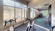 This well-appointed fitness studio is designed to support a comprehensive workout experience within a single, thoughtfully organized space. The room is finished with blue textured carpeting underfoot and neutral, subtly textured wall coverings that lend a clean, polished aesthetic throughout.

A dedicated cycling area anchors one end of the studio, equipped with True brand stationary bikes positioned to face a large wall-mounted flat-screen television, ideal for guided rides or entertainment during workouts. A wall-mounted control panel is conveniently located nearby.

Large floor-to-ceiling glass panel windows and a glass door connect the fitness studio to an adjacent lounge or common area, allowing natural light to flow generously into the space and creating a bright, open atmosphere. Clerestory windows above further enhance the sense of airiness.

Full-length wall mirrors on one side amplify the perceived depth of the room while providing functional visibility during exercise. Additional equipment is visible along the mirrored wall, including stability balls, exercise mats, and cardio machines, offering a variety of training options.

The studio is accessible through a standard interior door and features ceiling ventilation, ensuring a comfortable environment during use. Overall, the space presents a focused, amenity-quality fitness environment.