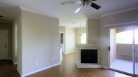 This single-story home offers a welcoming and functional layout with well-defined living spaces. The main living area is highlighted by a tile-surround fireplace with a raised hearth, serving as a natural focal point in the room. A ceiling fan with a light fixture overhead provides both ventilation and ambient lighting, while crown molding along the ceiling adds a refined architectural detail throughout the space.

The flooring throughout the main area consists of warm hardwood-style planks that contribute to a cohesive and inviting atmosphere. Neutral, light beige walls complement the natural tones of the floor, creating a bright and airy feel. A pendant light fixture is visible in the adjacent area, suggesting a defined dining or transitional space that connects to the rest of the home.

Beyond the main living area, a glimpse into what appears to be a laundry or utility room is visible, indicating practical amenities conveniently located within the home. A window allows natural light to filter into the adjacent space, enhancing the overall brightness. The clean, open layout of this single-floor home makes it an efficient and comfortable living environment.