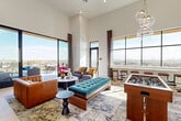 Located in the vibrant Heights neighborhood of Houston, TX, this elevated amenity lounge delivers a sophisticated social experience defined by dramatic architecture and sweeping city views. Inside, soaring double-height ceilings create an airy, expansive atmosphere, complemented by large floor-to-ceiling windows and glass doors that flood the space with abundant natural light. Light wood flooring runs throughout, anchoring an eclectic mix of furnishings that includes cognac leather seating, a tufted teal bench, and a bold gallery wall set against a deep teal accent feature. A generously sized wet bar with a white quartz countertop, bar seating, and a warm wood-paneled ceiling with pendant lighting serves as a central gathering point. A foosball table adds a playful, recreational touch. The space opens directly onto a wraparound outdoor terrace that showcases unobstructed panoramic views of the Houston skyline and the surrounding neighborhood rooftops. The terrace is thoughtfully appointed with multiple seating zones, high-top bar tables, a built-in fireplace, an outdoor television, and a wood-clad ceiling overhead — creating an ideal setting for both relaxed lounging and lively entertaining under the open sky.