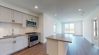 This contemporary single-floor residence offers a thoughtfully designed open-concept layout that seamlessly connects the kitchen and living areas. The kitchen is a standout feature, equipped with stainless steel appliances including a gas range, built-in microwave, dishwasher, and refrigerator. Shaker-style cabinetry in a soft grey tone is complemented by a classic white subway tile backsplash and a central island with a neutral countertop, providing ample workspace and storage. Warm hardwood-style flooring flows throughout the main living areas, creating a cohesive and inviting atmosphere. The spacious living area is bathed in natural light through large windows, offering pleasant outdoor views. A private balcony extends directly from one of the rooms, further enhancing the connection to the outdoors. The bedrooms feature plush carpeting and ceiling fans, with large windows allowing generous natural light. The primary bathroom is well-appointed with a double-sink vanity topped with crisp white quartz countertops, modern black hardware, a large mirror, and a glass-enclosed shower. A secondary bathroom offers a clean, minimalist design with an undermount sink and matching white cabinetry. Recessed lighting throughout lends a polished, modern finish to this well-appointed residence.