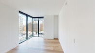 This well-appointed one-bedroom, one-bathroom residence combines modern design with functional living across a single floor. Light hardwood flooring flows consistently throughout the space, creating a cohesive and airy aesthetic.

The open-plan kitchen and living area form the heart of the home. The kitchen is thoughtfully designed with warm wood cabinetry extending to the ceiling, a textured tile backsplash, stone countertops, and a full suite of stainless steel appliances including a refrigerator, dishwasher, range, and microwave. The adjacent living area benefits from dramatic floor-to-ceiling corner windows framed in sleek black, flooding the space with natural light and offering expansive urban skyline views.

The bedroom continues the clean, minimalist aesthetic with the same hardwood flooring and its own set of large floor-to-ceiling windows showcasing city views. A closet and access to the hallway complete the room's practical layout.

The hallway connects the main living areas to the bedroom and bathroom, and features a louvered closet door adding a subtle architectural detail. The bathroom is finished with large-format neutral tiles surrounding a soaking tub and shower combination, complemented by modern brushed metal fixtures and a wood-toned vanity. The overall ambiance is clean, contemporary, and light-filled.