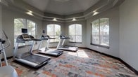 Located at 1017 N Shady River Ct in Benbrook, TX, this single-level fitness center is thoughtfully designed to accommodate a wide range of workout routines. The space is divided into distinct functional zones, each offering a different category of exercise equipment.

The cardio area is housed within an octagonal alcove defined by a tray ceiling and multiple large arched windows that frame views of the surrounding trees, filling the space with natural light. Three treadmills with individual monitors are positioned to take full advantage of the outdoor views, creating an inviting environment for cardio training.

The strength and free weights section features floor-to-ceiling mirrors that visually expand the room, a multi-station cable machine, an adjustable weight bench, and a full rack of dumbbells. A ceiling fan and recessed lighting keep the area comfortable and well-lit.

A dedicated functional training zone offers TRX suspension trainers, medicine balls, kettlebells, resistance bands, plyo boxes, and a spin bike, catering to high-intensity and cross-training workouts.

Throughout the facility, a bold geometric carpet in gray and orange tones adds a modern aesthetic, while decorative columns and dark crown molding contribute architectural character. The overall design balances practicality with an attractive, motivating atmosphere.