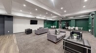 This well-appointed multi-use amenity space offers a generous, open floor plan thoughtfully divided into two functional zones. The main lounge area features plush gray sofas and sleek black armchairs arranged around a patterned carpet, creating a comfortable and inviting gathering environment. Multiple wall-mounted flat-screen TVs are positioned throughout for easy viewing from various seating arrangements. Recessed lighting set within a drop-tile ceiling illuminates the space evenly, while exposed silver ductwork along the perimeter adds an industrial design touch.

Transitioning toward the rear, the space shifts to a dedicated collaboration and meeting area finished with warm wood-look flooring. White rectangular tables paired with modern white chairs accommodate multiple small groups simultaneously, and a freestanding whiteboard serves as a focal point for presentations or brainstorming sessions. The walls in this zone are painted in a deep charcoal-gray, providing a focused, professional atmosphere.

A separate alcove, accented with bold green walls, provides additional flexible seating and table space alongside a freestanding bookshelf, and is accessible via two secured exit doors. The overall design balances modern aesthetics with practical functionality, making this space well-suited for social gatherings, meetings, and collaborative work sessions.