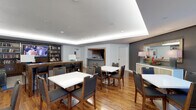 This single-floor communal space is thoughtfully designed to accommodate gathering, dining, and relaxation. The room features warm hardwood flooring throughout, complemented by a sophisticated tray ceiling with recessed LED cove lighting that lends a clean, modern aesthetic to the space. Multiple white-topped dining tables, each surrounded by wood-framed chairs with charcoal upholstered seats, are arranged to allow for both intimate and group settings. Along one wall, a built-in media unit houses a flat-screen television and a well-stocked bookshelf, creating a casual lounge-like atmosphere. A pass-through kitchenette is visible toward the back of the room, outfitted with cabinetry and counter space to support food and beverage service. A separate door nearby suggests additional utility or storage access. On the opposite side of the room, a decorative credenza topped with table lamps and a framed mirror adds an elegant finishing touch. The neutral gray and white color palette, accented by warm wood tones, creates a welcoming and versatile environment suitable for a variety of social functions.
