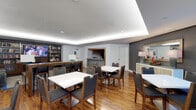 This single-floor communal space is thoughtfully designed to accommodate gathering, dining, and relaxation. The room features warm hardwood flooring throughout, complemented by a sophisticated tray ceiling with recessed LED cove lighting that lends a clean, modern aesthetic to the space. Multiple white-topped dining tables, each surrounded by wood-framed chairs with charcoal upholstered seats, are arranged to allow for both intimate and group settings. Along one wall, a built-in media unit houses a flat-screen television and a well-stocked bookshelf, creating a casual lounge-like atmosphere. A pass-through kitchenette is visible toward the back of the room, outfitted with cabinetry and counter space to support food and beverage service. A separate door nearby suggests additional utility or storage access. On the opposite side of the room, a decorative credenza topped with table lamps and a framed mirror adds an elegant finishing touch. The neutral gray and white color palette, accented by warm wood tones, creates a welcoming and versatile environment suitable for a variety of social functions.